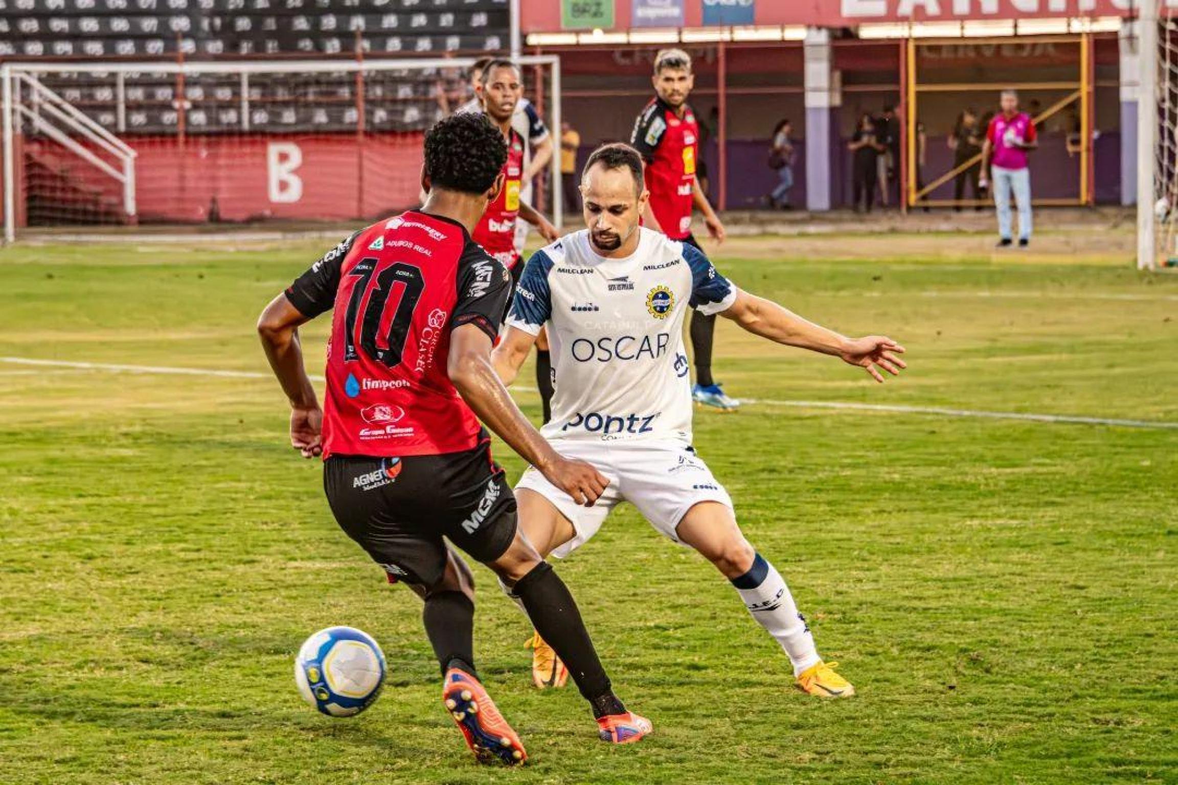 A imagem mostra uma partida de futebol entre o Pouso Alegre FC, de camisa vermelha, e o São José EC, de camisa branca. Um jogador do Pouso Alegre, com a camisa número 10, conduz a bola enquanto um jogador do São José se aproxima para marcá-lo. A partida parece estar acontecendo em um estádio, com arquibancadas e pessoas ao fundo.