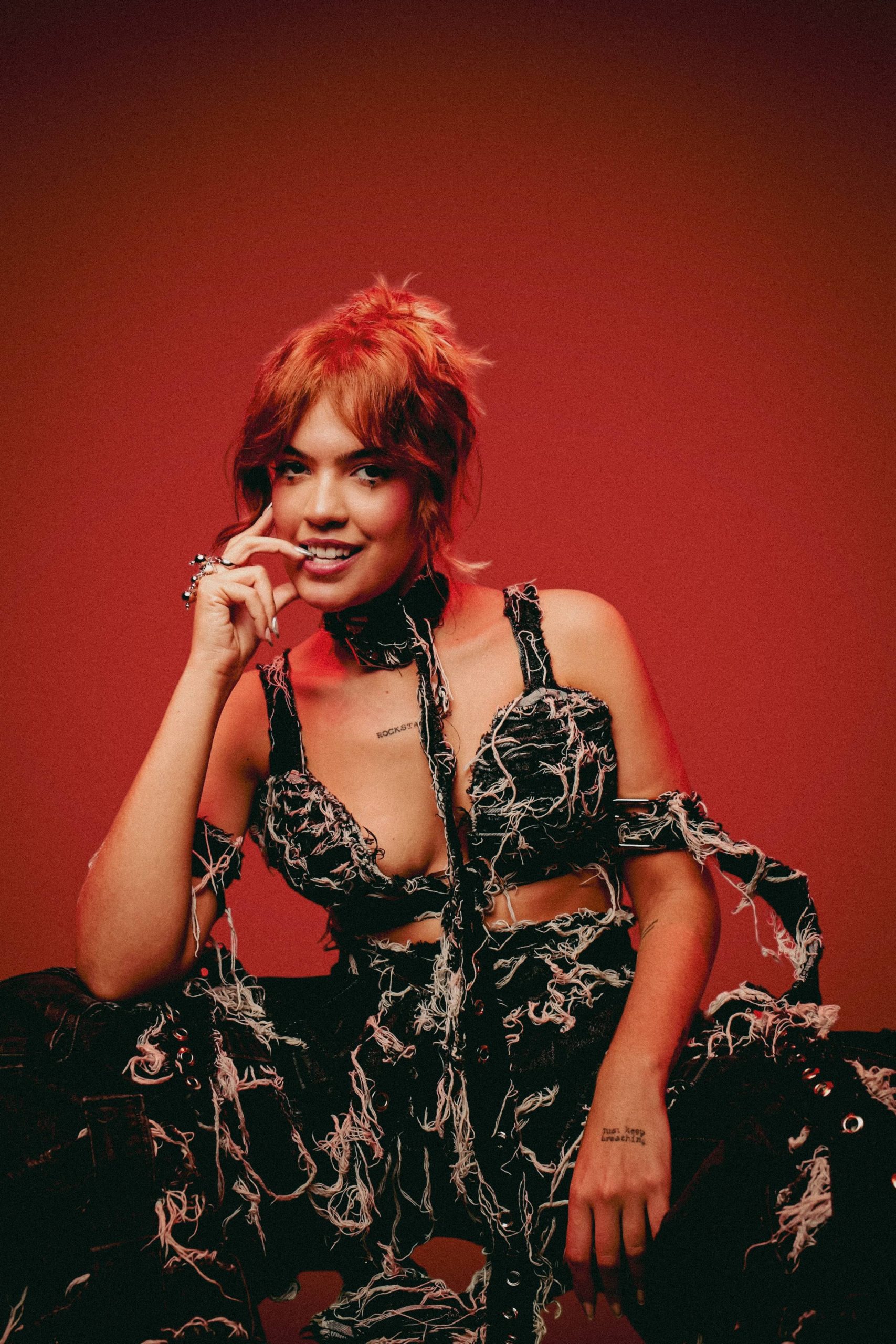 Cantora Blue com cabelos curtos avermelhados, vestindo conjunto preto com detalhes desfiados, posa sorridente em frente a um fundo vermelho.