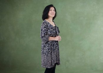 Yoko Shimomura, de cabelos escuros médios, vestindo um vestido estampado em tons de preto e bege, posa de perfil contra um fundo verde.