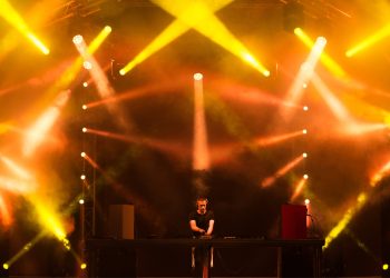 DJ se apresenta no palco com fortes luzes amarelas e efeitos visuais em um evento de música eletrônica.