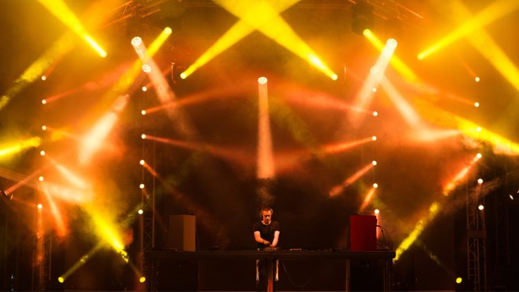 DJ se apresenta no palco com fortes luzes amarelas e efeitos visuais em um evento de música eletrônica.